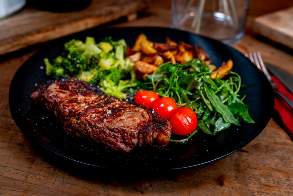 Ein Steak, Tomaten, Rucolasalat und weitere Beilagen auf einem dunklen Teller. Das Gericht steht auf einem Holztisch